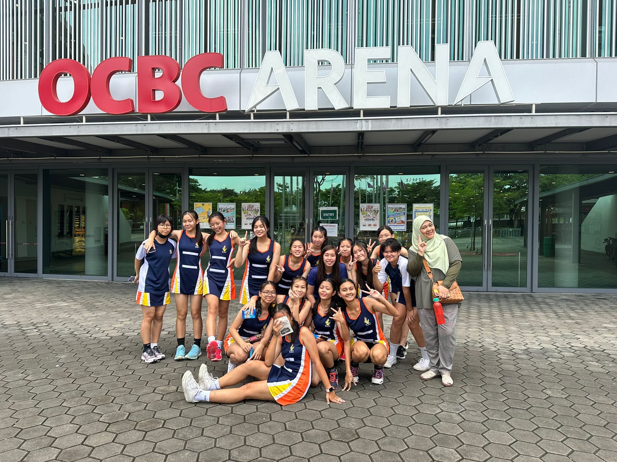 A group of young female athletes in navy and orange uniforms pose together outside the OCBC Arena.